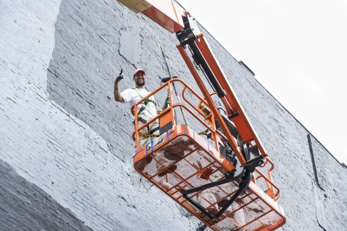 Painter Rafael Macia of Dorchester hangs over the bucket of his lift as he primes the wall on Washington Street for the Mural Festival